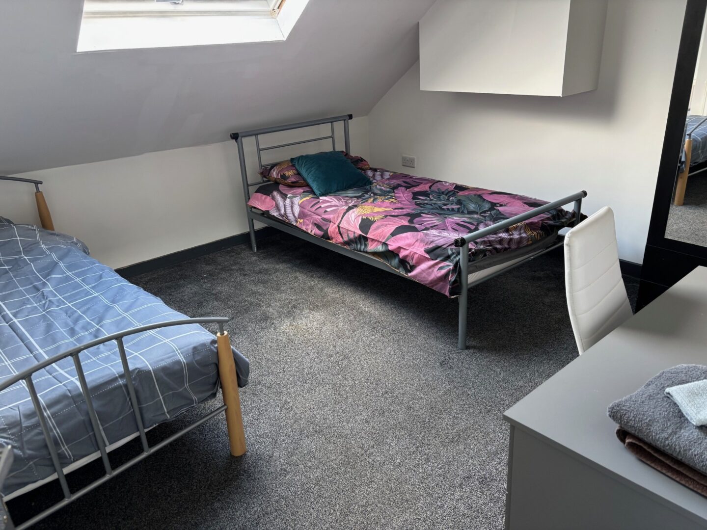 Attic bedroom with two single beds, floral and plaid bedding, skylight, desk, and chair on gray carpet.