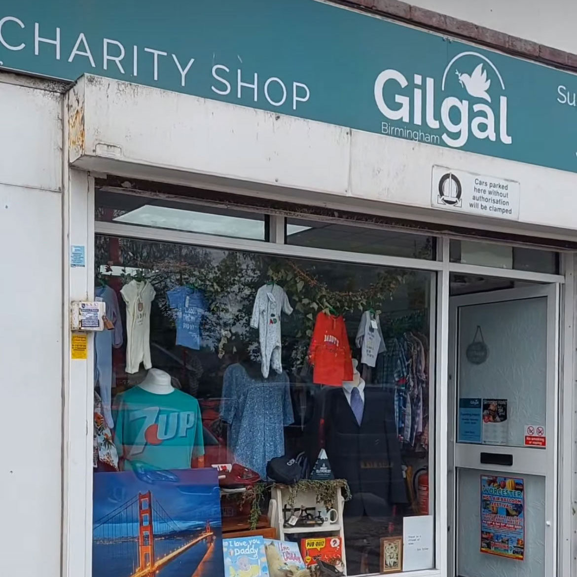 A charity shop window displays a variety of clothing items, including baby onesies, shirts, and a green "7UP" t-shirt. There are also books and a framed picture of the Golden Gate Bridge visible in the display. The shop sign reads "Gilgal Charity Shop" and indicates its location in Birmingham.