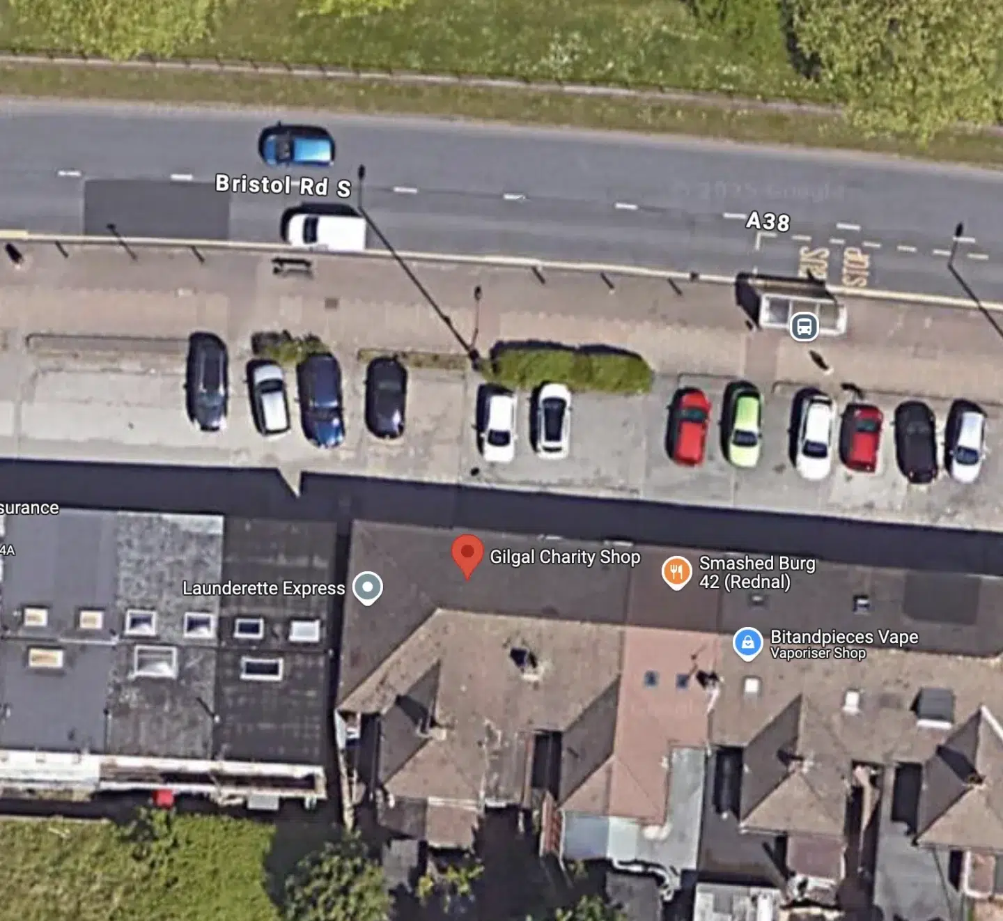 Aerial view of a street with parked cars along one side, featuring several businesses including a charity shop, a laundrette, and a vape shop. The road is labeled as Bristol Rd S, with a bus stop indicated nearby.