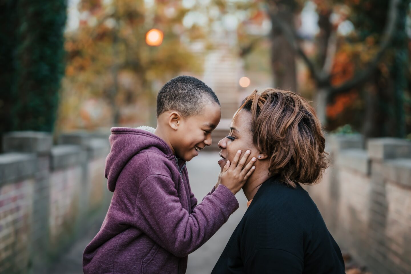 A young boy in a purple hoodie smiles and touches the face of a woman with short, curly hair, both sharing a joyful moment outdoors. The background features blurred trees with autumn foliage.