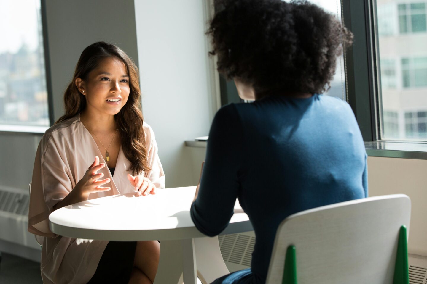 Two women are seated at a round table in a well-lit office space. One woman, wearing a light pink top, is animatedly speaking and gesturing, while the other woman, dressed in a dark blue top, is facing her and listening attentively.