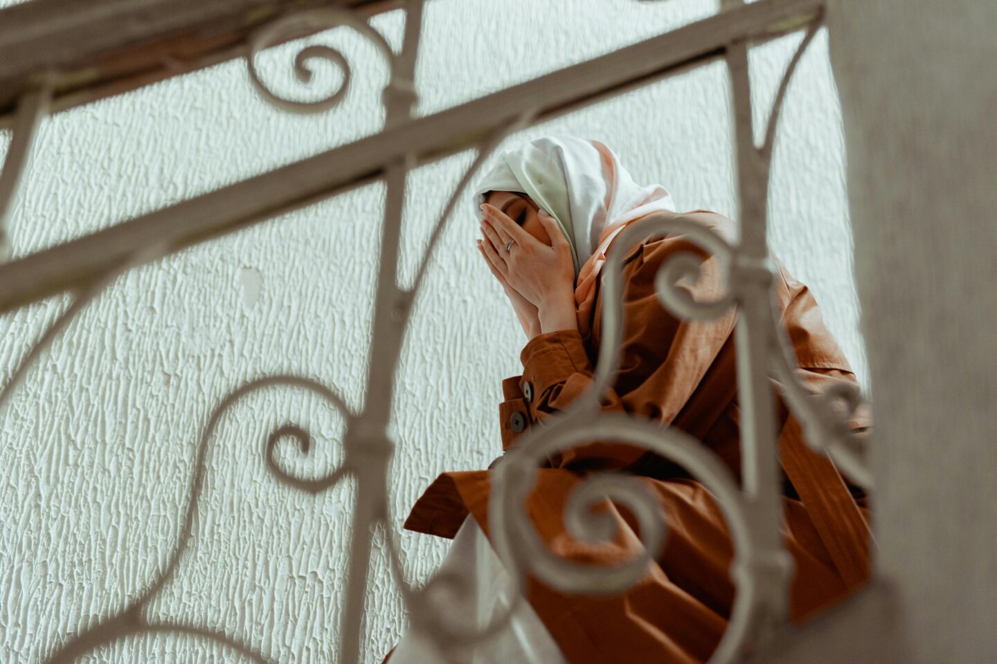 A woman wearing a headscarf and a brown coat is sitting on a staircase, covering her face with her hands. The background features a textured wall and ornate railing.